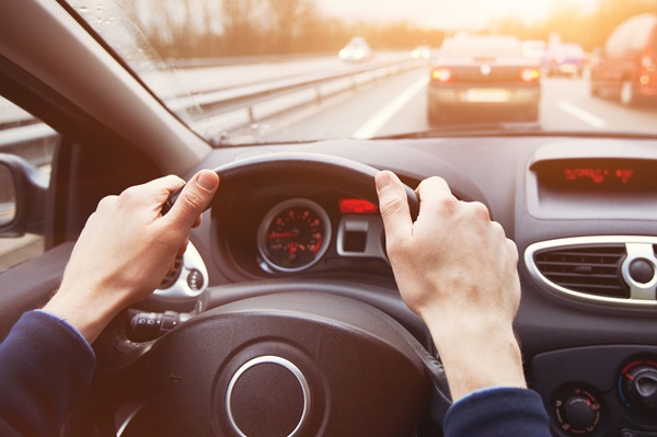 Driver’s view of hands on a steering wheel while traveling in the left lane of a multi-lane highway with traffic ahead.