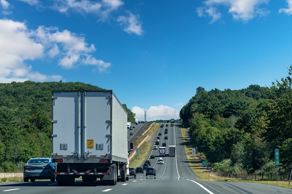 Semi-truck traveling in the right lane of a divided highway with multiple vehicles ahead under a partly cloudy sky.