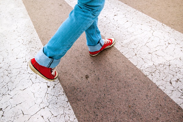 Close-up of a person wearing red sneakers and jeans walking across a cracked crosswalk on a city street.