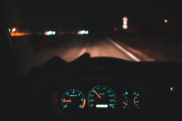 View from inside a car at night showing the illuminated dashboard and speedometer, with the road ahead visible through the windshield and headlights blurring in the distance.