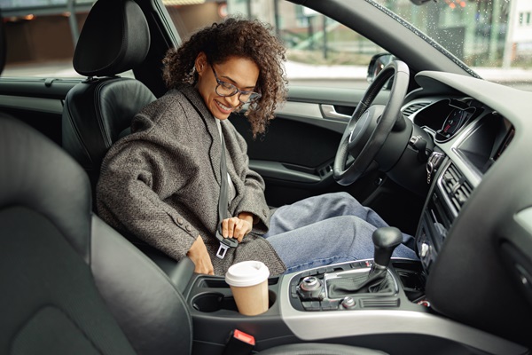 A woman with curly hair and glasses sitting in the driver's seat of a car, smiling as she fastens her seatbelt before driving.