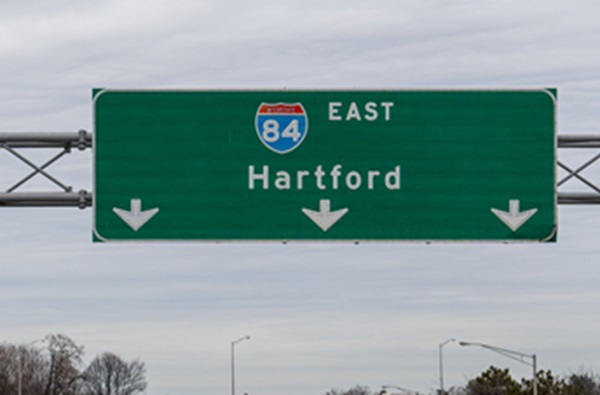 A large green highway overhead sign for Interstate 84 East toward Hartford, featuring three white down-pointing arrows indicating lane alignment, set against a cloudy overcast sky.