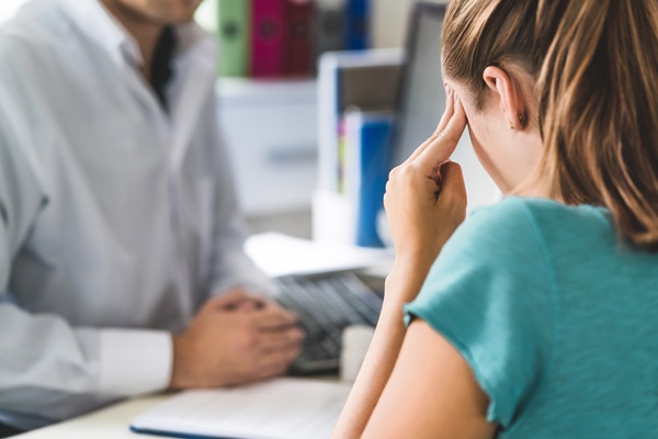 A woman rubbing her temples during a medical consultation with a doctor, symbolizing the diagnosis and discussion of traumatic brain injury symptoms.